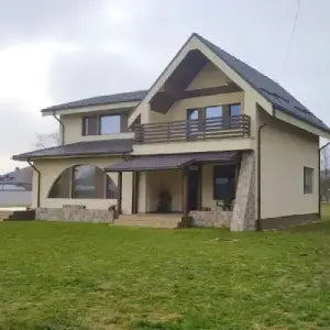 A modern two-story house with beige walls and a dark gray roof, featuring large windows and a balcony. It's set on a green lawn under a cloudy sky.